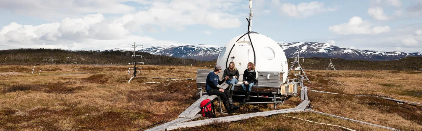 Researchers sitting beside a field monitoring station in an open tundra landscape