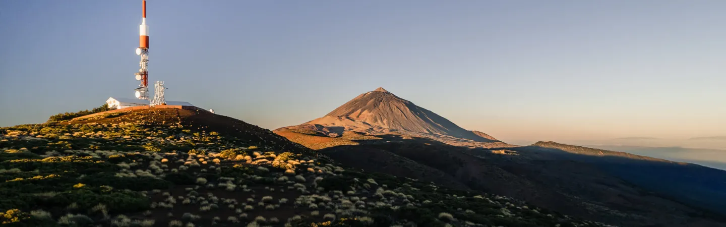 Atmospheric measurement tower on hillside with volcanic mountain in the background at sunset.