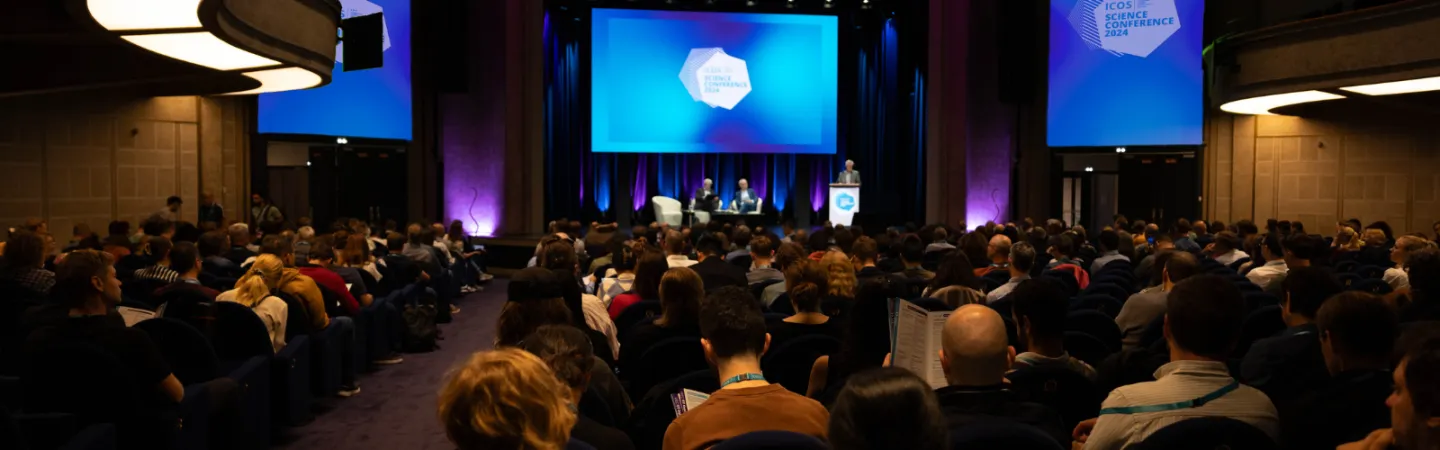 A large auditorium filled with seated attendees faces a stage where a speaker stands at a podium and three panelists sit beside them. The stage is lit with purple and blue lighting, and a large screen behind the presenters displays the text “ICOS Science Conference 2024.”